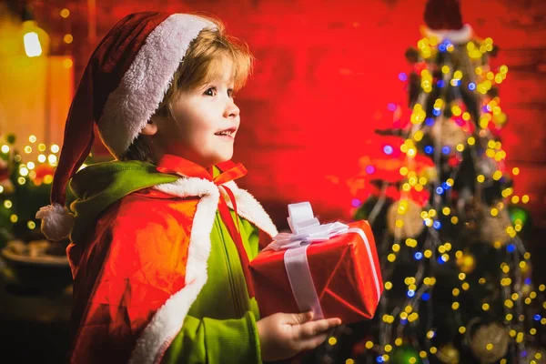 Menino feliz perto da árvore de Natal com presente de Natal. O miúdo está a usar roupa de Pai Natal. Conceito de crianças Natal. — Fotografia de Stock