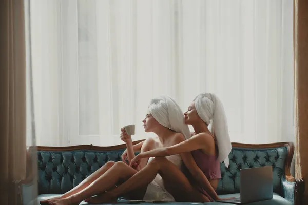 Attractive girls having breakfast in bed with laptop in modern hotel. Joyful female friends lying in bed at living room. — Stock Photo, Image