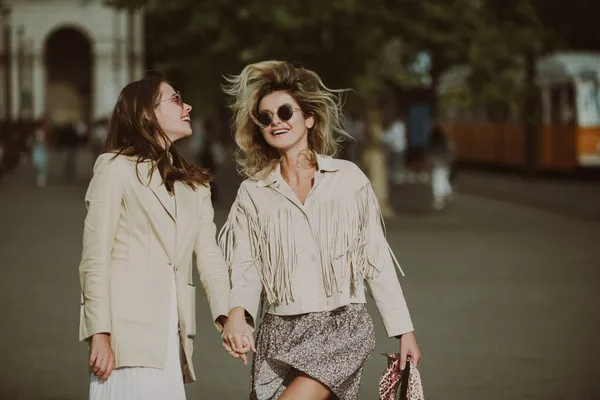 Dos mujeres emocionadas caminando por la calle. Hermosa chica cogida de la mano con un amigo mientras camina por la ciudad. Retrato al aire libre de dos hermanas riendo pasando tiempo libre juntas. — Foto de Stock