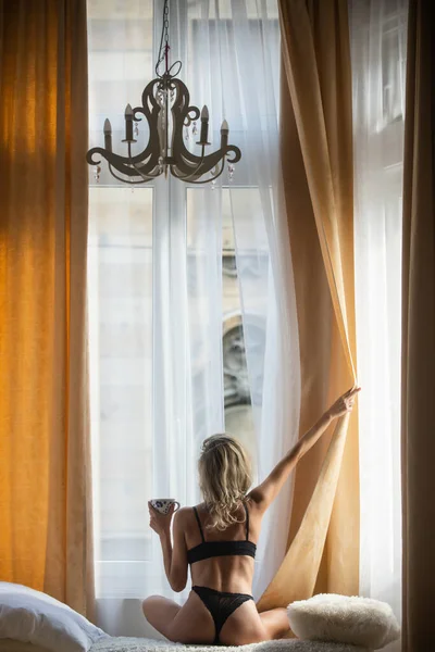 Πίσω όψη των νέων ταιριάζει υγιείς γυναίκες στο υπνοδωμάτιο μετά ξυπνήσει. Πρωινό πίνοντας καφέ. Γυναίκα ανοίγει κουρτίνες παραθύρων. Οπίσθια όψη σε σέξι γυναίκα σε πολυτελές διαμέρισμα ή δωμάτιο ξενοδοχείου. — Φωτογραφία Αρχείου