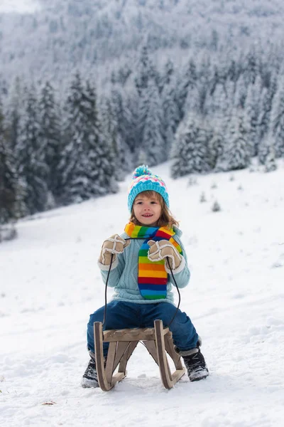 Porträt eines glücklichen kleinen Kindes mit Strickmütze, Schal und Pullover. Junge genießt eine Schlittenfahrt. Kinderschlittenfahren im Schnee. Winterliche Weihnachtslandschaft mit Schnee. — Stockfoto