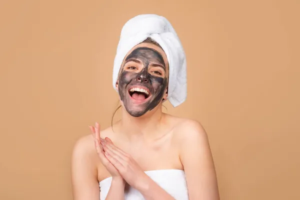 Lera ansiktsmask, ansiktsmask spa. Vacker skrattande kvinna med kosmetisk lera ansiktsbehandling, spa hälsa koncept. Skönhetsbehandling av huden. Handduk på huvudet. Beige bakgrund, isolerad. — Stockfoto