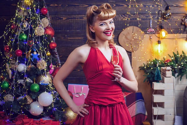 저녁에 붉은 드레스를 입은 여성 이 크리스마스 트리 배경에 반짝 이는 와인을 곁들여 입고 있다. 크리스마스에 유행하는 패션 메이크업 과 헤어 스타일. — 스톡 사진