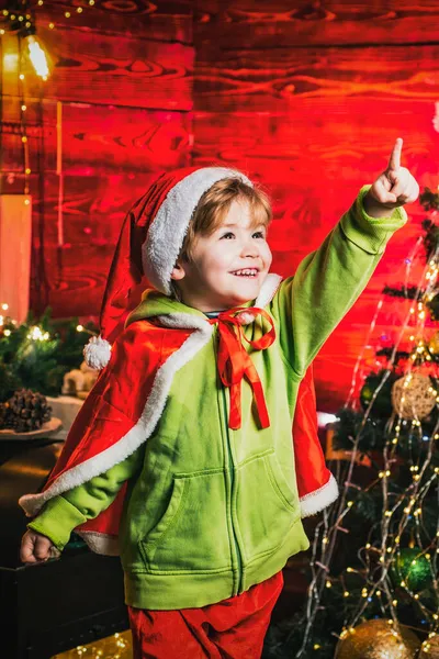 Çocuklar için sihirli Noel gecesi. Noel ağacının yanındaki parmağıyla mutlu, heyecanlı, gülümseyen çocuk. Küçük çocuk Noel Baba kıyafeti giyiyor.. — Stok fotoğraf