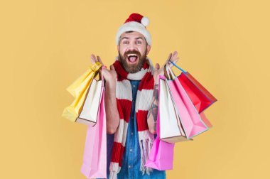 Noel alışverişçisi Noel Baba şapkası ve eşarbıyla gülümsüyor. Adam sarı arka planda alışveriş torbaları tutuyor. Noel 'de Adam