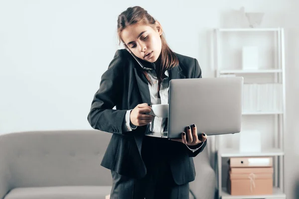 Empresaria sentarse en el interior de la oficina utilizando ordenador portátil en la oficina. Trabajador de oficina ocupado bebiendo café, hablando por teléfono, usando computadora portátil. — Foto de Stock