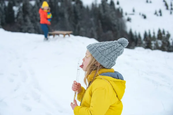 Vtipné vánoční děti olizují sněžný rampouch jazykem. Dětská koncepce nachlazení a chřipky. Děti v teplém zimním oblečení ve sněhovém počasí. Šťastné dítě dívka hraje na zimní procházku v přírodě. — Stock fotografie