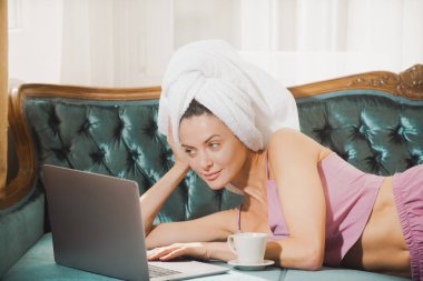 Evde pijama ve havlu giyen, dizüstü bilgisayar kullanan, kahve içen çekici bir genç kız..