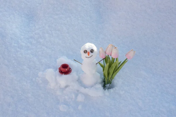 Χιονάνθρωπος με φλιτζάνι καφέ και ανοιξιάτικα λουλούδια τουλίπες. Χαρούμενος χαμογελαστός χιονάνθρωπος την ηλιόλουστη μέρα του χειμώνα. — Φωτογραφία Αρχείου