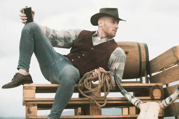 Cowboy on ranch. Handsome man in cowboy hat and retro vintage outfit. Guy drinking whiskey. — Stock Photo, Image