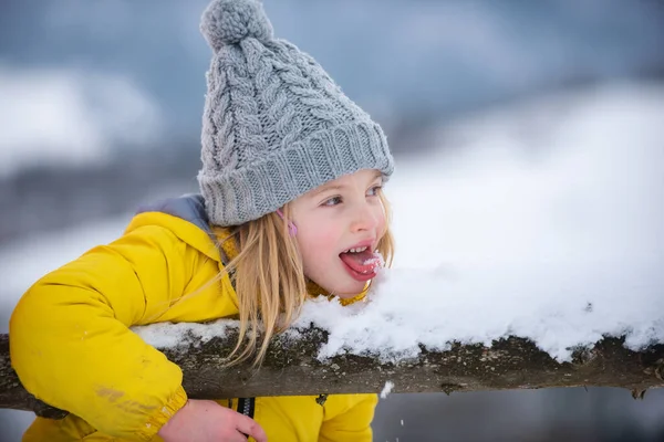 Vinterflicka äter snö utomhus. Roliga julungar slickar snö med tunga. Koncept för förkylning och influensa. Barn som bär varma vinterkläder i snöväder. — Stockfoto