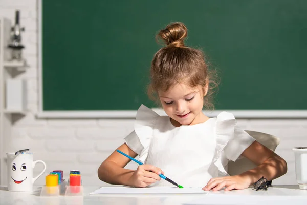 Dziecko dziewczyna rysuje w klasie siedzi przy stole, bawiąc się na tle tablicy szkolnej. dziecko ucznia nauka malarstwa. — Zdjęcie stockowe