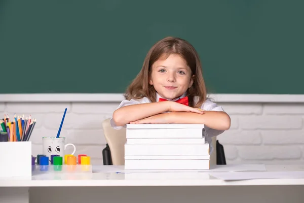 Leende flicka sitter vid skrivbordet och håller händerna på böckerna i skolans klassrum. Grundskola och grundskola. — Stockfoto