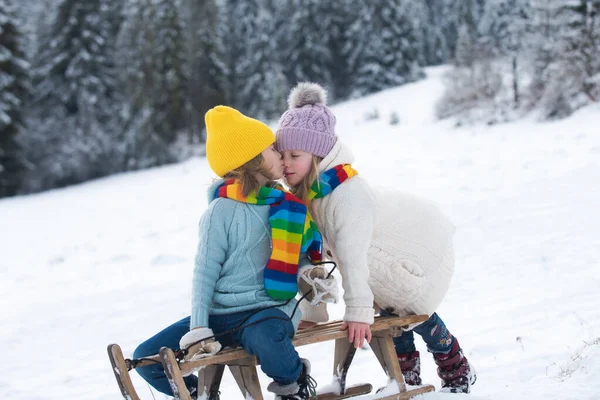 Les enfants s'embrassent. Joyeux petit garçon et petite fille sur luge en hiver. Les enfants aiment monter sur des glissades de neige en hiver. Enfants garçon et fille profiter d'une promenade en traîneau et baisers. — Photo