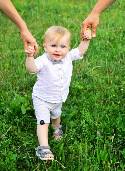 Los padres de la mano y el niño primer paso. Bebé jugando en hierba verde. Niño divirtiéndose en picnic familiar en el jardín de verano. Retrato de una familia joven y feliz enseñando al bebé a caminar en el parque. —  Fotos de Stock