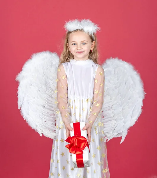 Kind engel klein meisje met cadeau, studio portret. Een engeltje met witte vleugels heeft een gave. Valentijnskaart. Schattig engelenmeisje met engelenvleugels, geïsoleerd op rood. Valentijnsdag. — Stockfoto