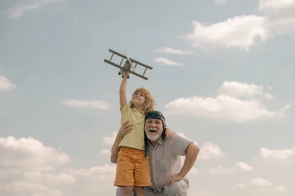 Kind jongen en grootvader met speelgoed vliegtuig over blauwe lucht en wolken achtergrond. Mannen generatie grootvader en kleinzoon spelen buiten. — Stockfoto