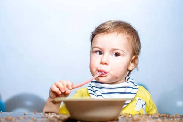 Baby eet kindervoedsel. Grappige jongen met bord en lepel. — Stockfoto