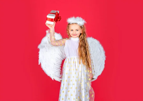 Kind engel klein meisje met cadeau, studio portret. Kleine engel, gelukkige fee met witte vleugels heeft een geschenk. Schattig engelenmeisje met engelenvleugels, geïsoleerd op rood. Valentijnsdag. — Stockfoto