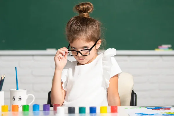 Gyermek lány rajz színező tollak festék. Aranyos kislány portréja, aki boldogan mosolyog, miközben élvezi a művészetet és a kézműves leckét az iskolában. Vicces iskolás lány arc szemüveggel. — Stock Fotó