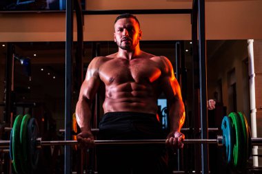 Halter kaldıran kaslı adam. Dambılı tişörtü olmayan atletik bir adamın portresi. Gövdesi kaslı, tişörtsüz bir adam spor salonunda..
