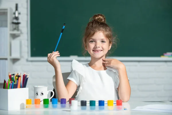 Kreatywny rozwój dzieci. Dziecko dziewczyna rysuje w klasie siedzi przy stole, bawiąc się na tle tablicy szkolnej. — Zdjęcie stockowe