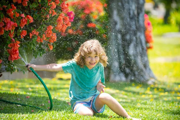 Happy kid boy pours water from a hose. Child watering flowers in garden. Home gardening. — Stock Photo, Image