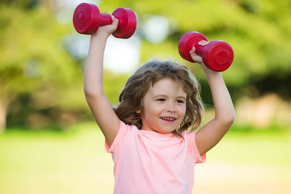 Glücklicher kleiner aufgeregter Junge, der im grünen Park Übungen mit Hanteln macht. Nahaufnahme Porträt eines sportlich lächelnden Kindes mit Hanteln. Glücklicher Junge beim Sport im Freien. — Stockfoto