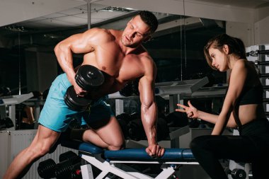 Kas yığını olan bir adamla kadın antrenör, kaslarını çalıştırıyor, dambıl kaldırıyor. Spor yapan atletik erkek sporcu. Spor konsepti. Sportif çift..