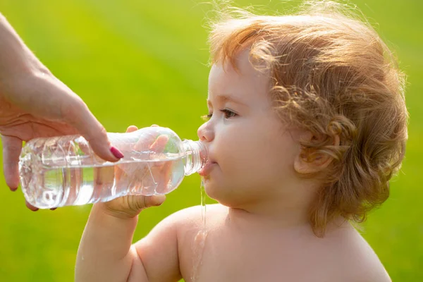 赤ん坊は母親の手から水を飲む。公園の中に巻きブロンドの髪の飲料水と赤ちゃんの男の子,ペットボトルを保持,屋外. — ストック写真