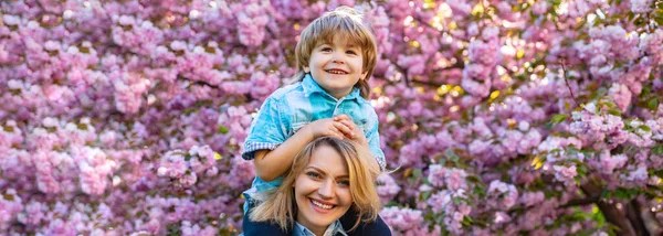 Bandiera primavera. Buona festa della mamma. Madre cavalcare figlio sulle spalle in giardino sakura. — Foto Stock