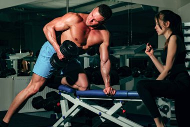 Sportif bir çift. Kas yığını olan bir adamla kadın antrenör, kaslarını çalıştırıyor, dambıl kaldırıyor. Sporcu ağırlık egzersizi yapıyor..