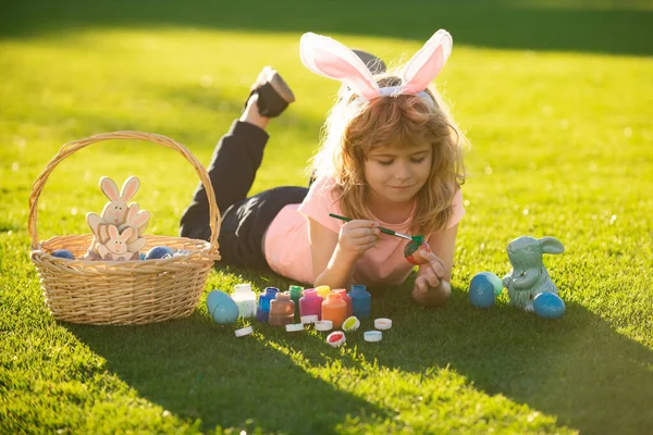 Pasen kinderen jongen in konijnenoren schilderen paaseieren buiten. Schattig kind in konijn kostuum met konijn oren hebben plezier in het park. — Stockfoto