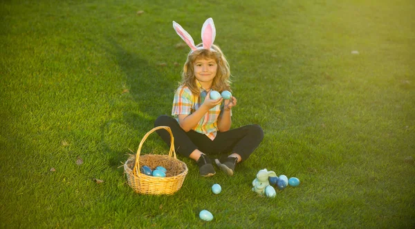 Kind met paaseieren in mand buiten. Jongen op gras in het park. Paaseieren zoeken. Fynny kinderen portret. Bunny kids met konijnenoren. — Stockfoto