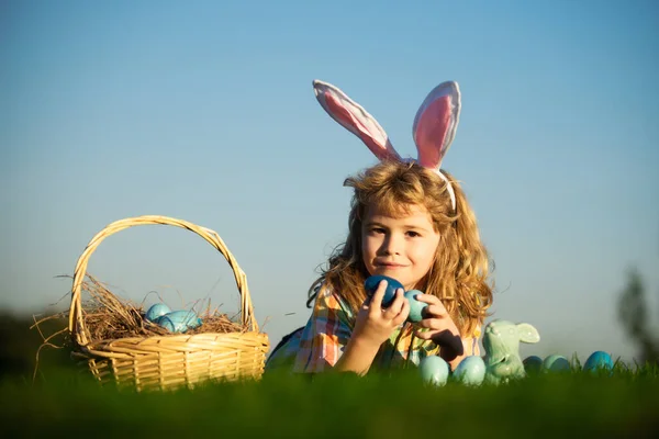 Child hunting easter eggs in backyard laying on green grass on sky background with copy space. — Zdjęcie stockowe