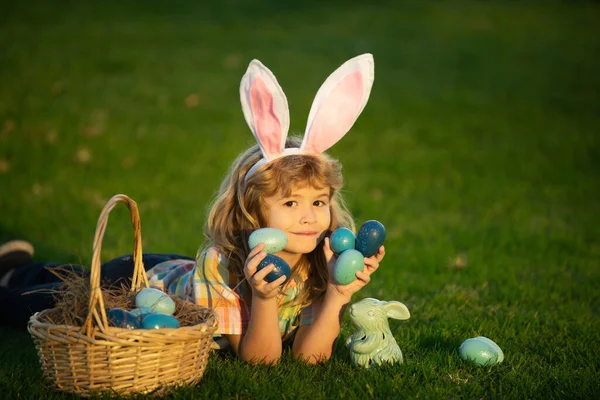 Paskalya yumurtalarını sepete koyan çocuk. Çocuk parkta çimlerin üzerinde yatıyor. Paskalya yumurtası avı. Fynny çocuk suratı. Tavşan kulaklı çocuk.. — Stok fotoğraf