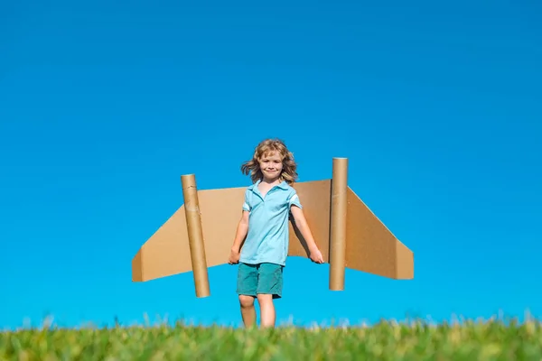 Niño feliz jugar con alas de cartón plano de juguete contra el cielo azul. Niño divirtiéndose en el campo de verano al aire libre. Retrato de niño con alas de papel. — Foto de Stock