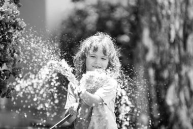 Yaz günü bahçede çiçekleri sulayan sevimli küçük bir çocuk. Çocuk bahçe hortumu kullanıyor. Bahçede çiçek sulayan komik çocuk..