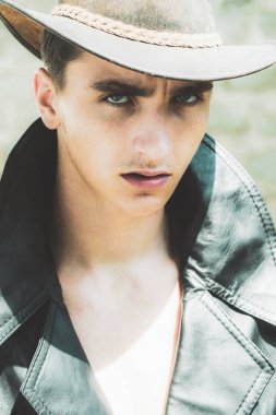 Tuğla duvarın önünde dikilip kameraya bakan kareli gömlekli genç adam..