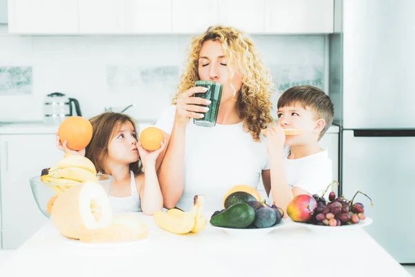 Мати і діти дочка і син п'ють зелену смужку на кухні з фруктами і овочами. Щаслива сім'я п'є зелену спіруліну на білій кухні . — стокове фото