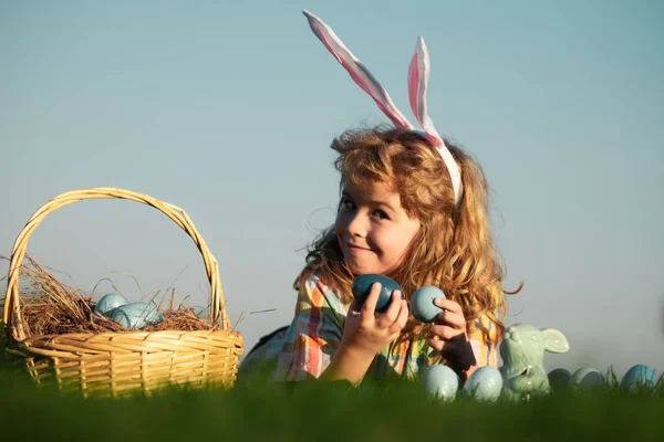 Children hunting easter eggs. Kid boy lying on the grass and finding easter eggs, on sky background with copy space. Child with easter eggs and bunny ears, outdoor portrait. — Zdjęcie stockowe