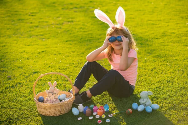 Child boy with bunny ears cover eyes with eggs on grass background. — Zdjęcie stockowe