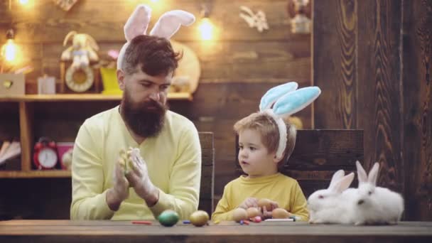 Vader en kind zoon die thuis paaseieren tekenen, eieren beschilderen. Papa en kind vieren Pasen met grappige konijnen. Ouders met kind in konijnenoren. — Stockvideo