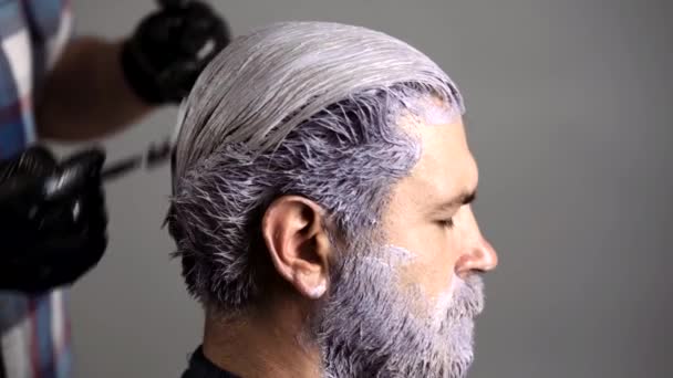 Cabeleireiro, homem de coloração capilar. Barbeiro sênior atraente fazendo um corte de cabelo e penteado para o cliente na barbearia. Cabeleireiro colorista tinge o cabelo de um homem barbudo. — Vídeo de Stock