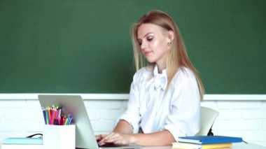 Masanın yanında dizüstü bilgisayarlı, gülen surat ifadesi olan tatlı bir kız. Genç öğretmen. Sınıftaki sevimli sarışın genç bayan öğretmen. Öğrenme ve eğitim kavramı. Öğretmenler Günü.