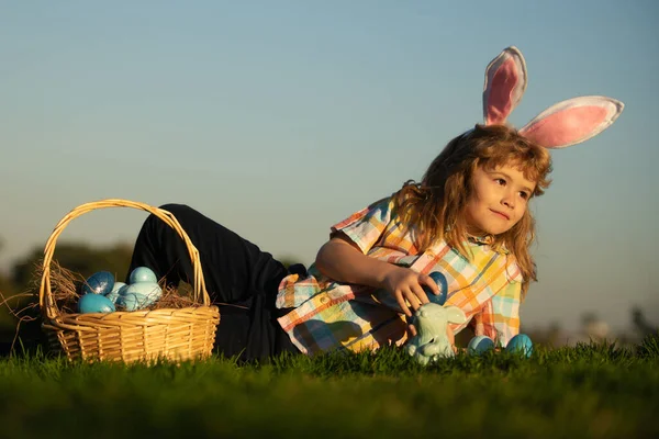 Cute bunny child boy with rabbit ears. Kids hunting easter eggs in park laying on grass on sky background with copy space. — стоковое фото
