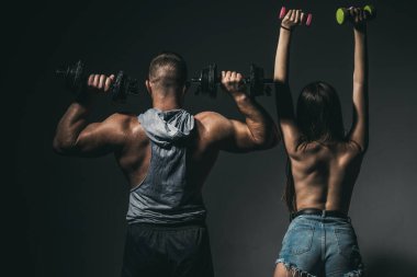Dambılla egzersiz yapan seksi spor çifti. Çıplak vücutlu kaslı bir adam, arka planda koyu renkli halterleri olan fitness kadını. Sıska seksi kız güçlü kaslı adam egzersizi yapıyor..