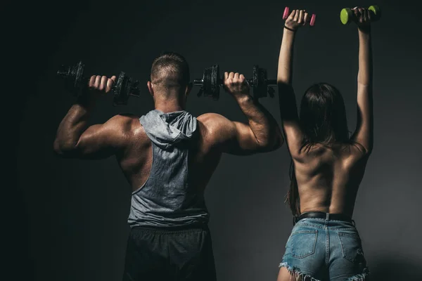 Sexy pareja deportiva haciendo ejercicio con mancuerna. Hombre musculoso con cuerpo desnudo, mujer fitness con mancuernas sobre un fondo oscuro, vista trasera. Slim sexy chica con fuerte muscular hombre entrenamiento. —  Fotos de Stock