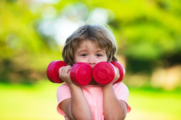 Cute funny little boy doing exercises with dumbbells in green park. Closeup portrait of sporty child with dumbbells. Happy child boy exercising outdoor. Healthy activities kids lifestyle. — Foto Stock