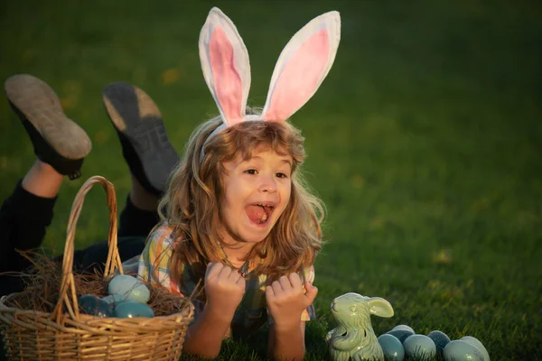 Child bunny boy with rabbit bunny ears. Child with easter eggs in basket outdoor. Boy laying on grass in park. Easter egg hunt. Fynny kids portrait. — Zdjęcie stockowe